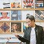 Makoto in a black leather jacket and gray t-shirt stands in a record shop aisle, holding and inspecting a vinyl album. Behind him, wooden shelves display rows of his previous album covers neatly arranged on the wall.