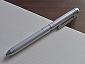 A sleek, cylindrical all-metal pen in matte silver finish, laid diagonally on an open grid notebook, featuring a clip and twist mechanism at the top, resting on a wooden table.