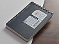A small, black spiral-bound memo pad with squared grid paper, showing its front cover label featuring date markings and a barcode, resting on a wooden table.