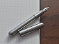 A compact stainless-steel fountain pen with its cap removed and placed next to it, revealing a conical nib and slim barrel, resting on a wooden table.